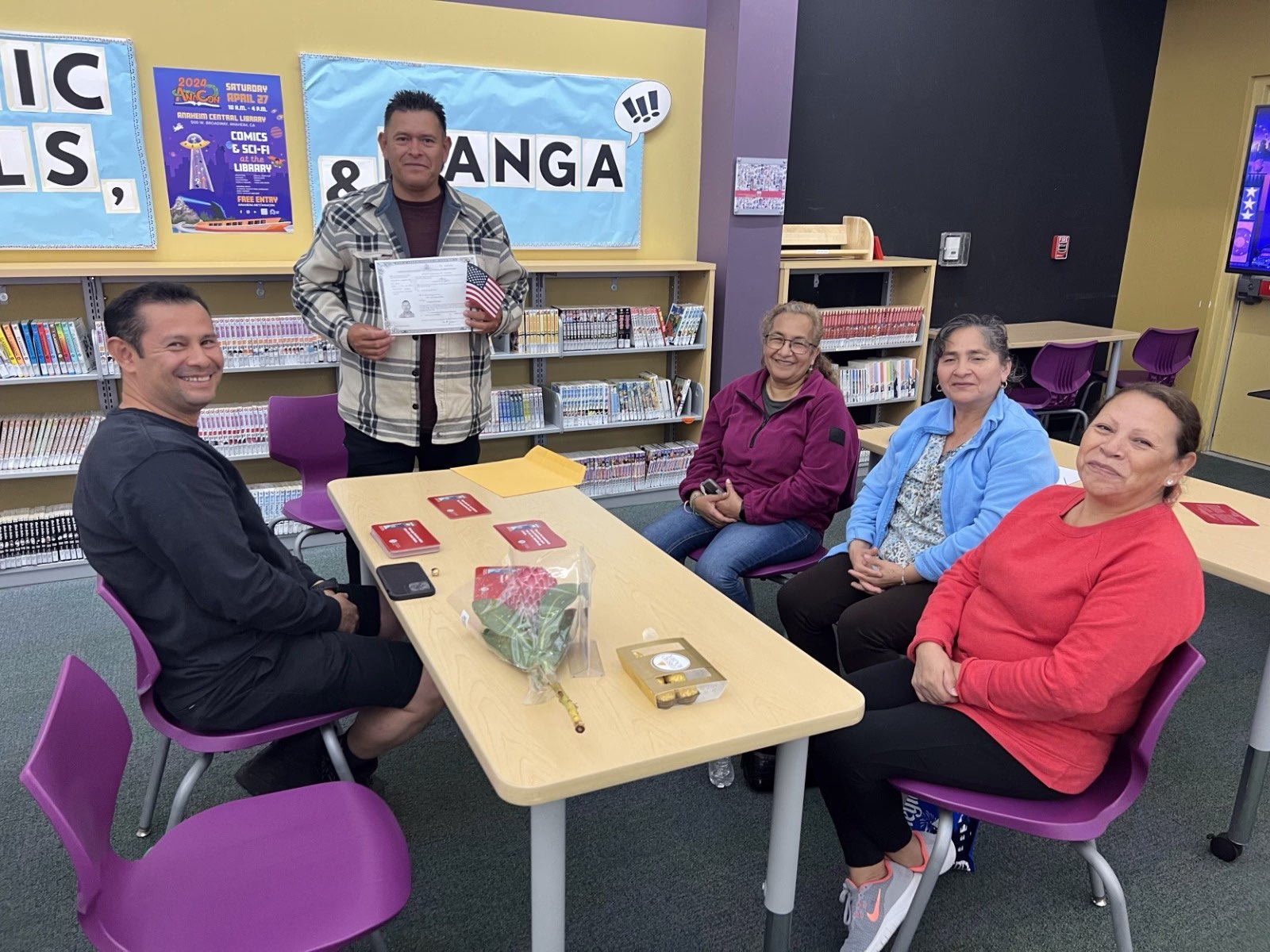 Five adult students sit around a table in a classroom/library setting during a citizenship class, smiling at the camera. One man stands behind them holding a certificate and a small U.S. flag, celebrating a milestone. On the table are red booklets, a bouquet of flowers, and a small box of chocolates. Bookshelves and educational posters are visible in the background.